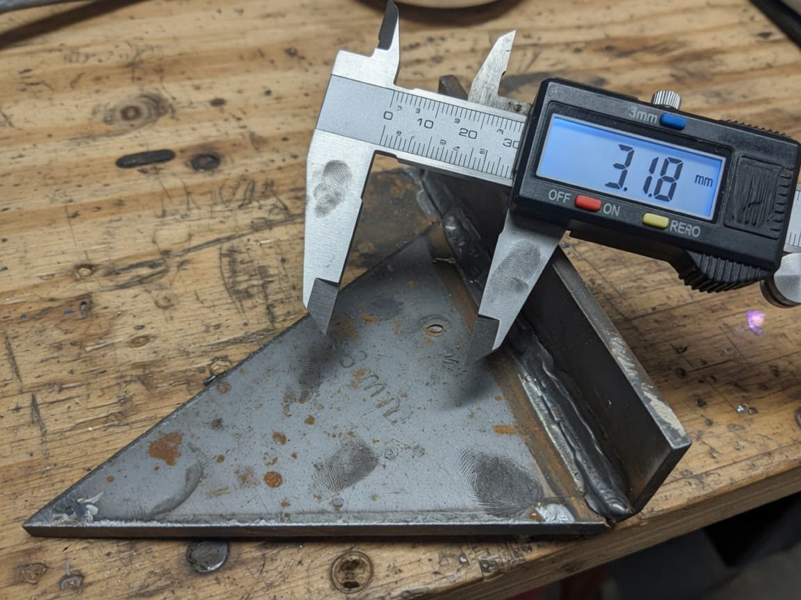 Digital caliper measuring a welded steel gusset on a worn wooden bench.