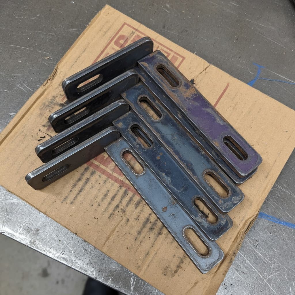 Short stack of mill-finish steel L-brackets with slotted holes on cardboard.